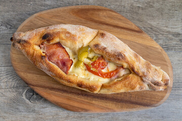 Peinirli with cheese, ham, tomato and green peppers baked and ready to serve. Greek open faced pizza on wooden cutting board and wooden kitchen surface.