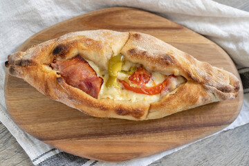 Peinirli with cheese, ham, and vegetables baked and ready to serve. Greek open faced pizza on wooden cutting board placed above kitchen towel.