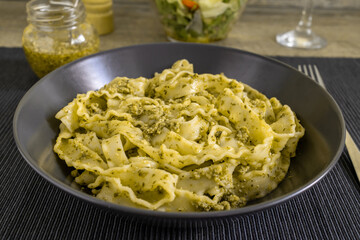 Pasta Mafaldine with tofu pesto sauce served on a dark plate. Salad bowl, glass jar with pesto sauce on textile placemat and wooden kitchen surface.