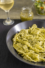 Pasta Mafaldine with tofu pesto sauce served on a dark plate. Salad bowl, glass jar with pesto sauce, a glass of wine on textile placemat and wooden kitchen surface.