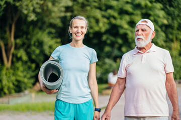 Fototapeta premium Active senior man and woman physiotherapist carrying exercise mats after sports workout in nature. Old man and his adult daughter holding yoga mats, walking along park path together, talking.