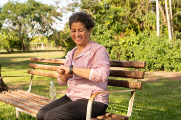 Smiling Elderly brazilian woman using smartwatch. Outside in park and woods. Technology, healthy,...