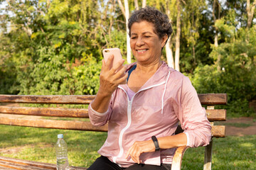 Senior black brazilian woman using smart cell phone and smiling. Outside in park and woods. E-commerce, online shopping, communication concept.