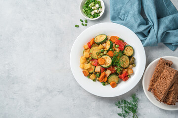 Baked zucchini, potatoes, carrots, peas and tomatoes with fresh herbs on a light gray background.
