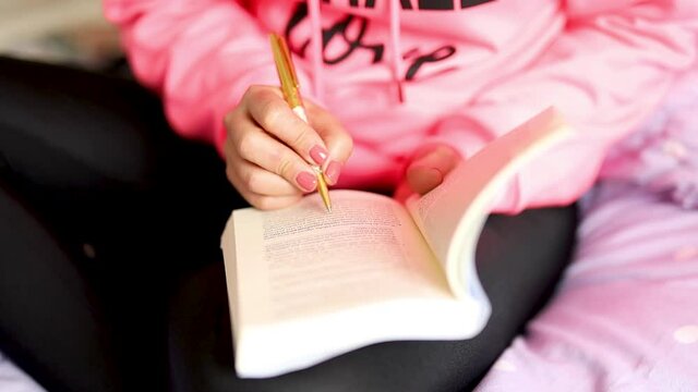 mujer con polo rosado sentada en la cama de su habitacion, con un libro estudiando feliz