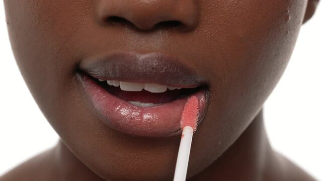 Close up of an african woman's mouth, applying lip gloss on her top and bottom lips. White background.