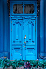 Ornate double door painted in bright blue color. Vintage wooden doorway and defocused faux conifer Christmas decoration on foreground. Classical architecture details