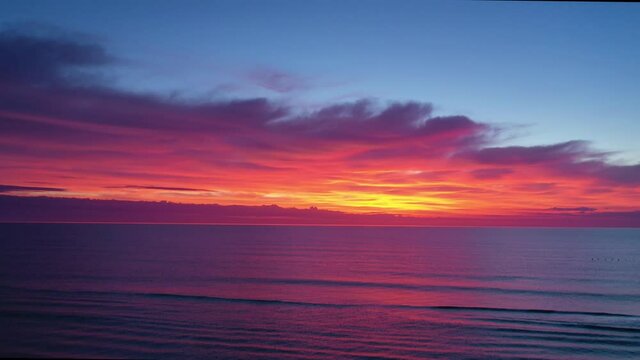 Tilt down aerial of dramatic predawn sky with colors of red, orange, yellow, and purple over a magenta and dark purple ocean.