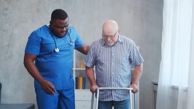 African-American caregiver is teaching disabled old man to walk with walker. Professional nurse and handicapped patient in a nursing home. Assistance, rehabilitation and health care.