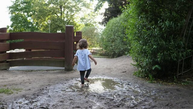 Little child girl enjoy walk and run, jump and make splashes on dirt and mud puddle water on park ground