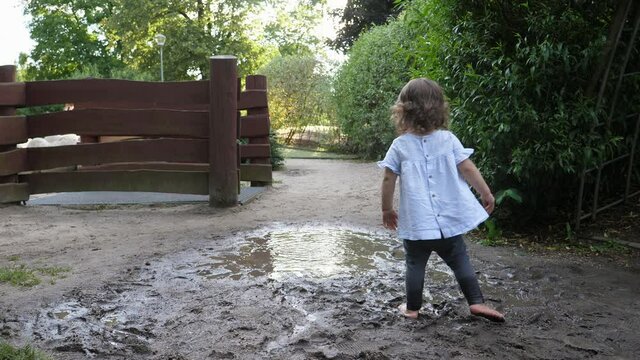 Little child girl enjoy walk and run, jump and make splashes on dirt and mud puddle water on park ground