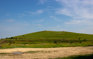 Fototapeta premium A green hill and a dirt road on the background of a blue sky with clouds