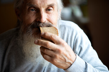 Old sad man with a long gray beard sitting by the table and eating soup and bread