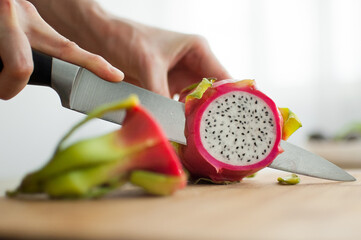 Female hands is cutting a dragon fruit or pitaya with pink skin and white pulp with black seeds on wooden cut board on the table. Exotic fruits, healthy eating concept