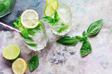 Glasses of basil lemonade with mint, basil, lime, lemon and ice on light gray background. Top view