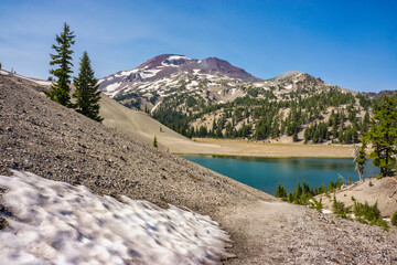 Patch of melting snow and a clear blue alpine lake surrounded by pumice covered moraine, fir trees,...