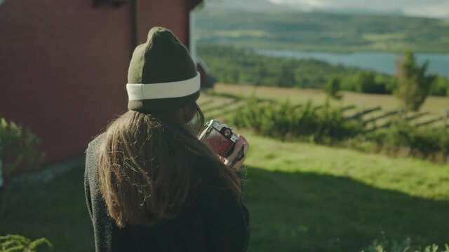 Authentic moment of a hipster in cozy relaxed clothes taking picture of the mountain lake view at her getaway to a classic red wooden cabin surrounded by green grass, concept of freedom, hygge, rest.