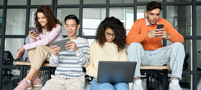 Young diverse multiracial cool group of high school college students girls and guys sitting at university campus space holding technology gadgets using digital devices studying together.