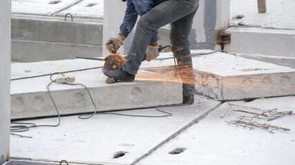 A professional builder cuts iron rebar with an angle grinder machine on a reinforced concrete slab at a construction site. Sparks fly from an electric circular saw. Construction of a new building.