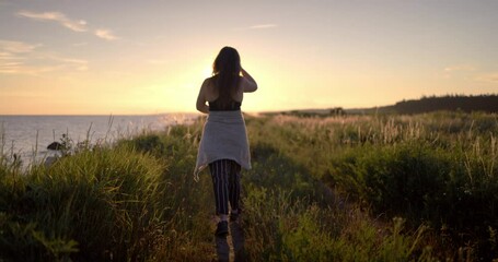 Girl walking in slow motion in the countryside in the golden light with backlight along a hiking trail by the coast and ocean - Powered by Adobe