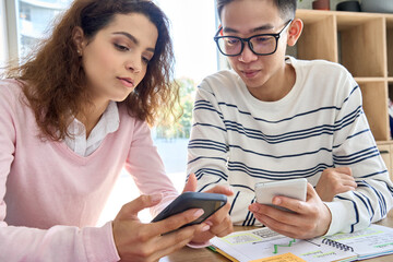 Gen z teens Asian guy and latin Hispanic girl using mobile cell phone social media applications sitting at desk in classroom. Diverse high school students holding smartphone learning in college. - obrazy, fototapety, plakaty