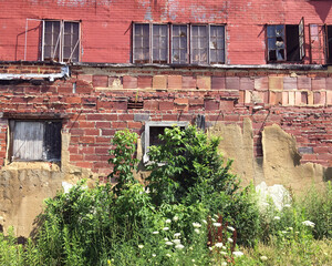 Abandoned building of red brick, fake brick finish and broken windows framed by overgrown weeds in the American Midwest.