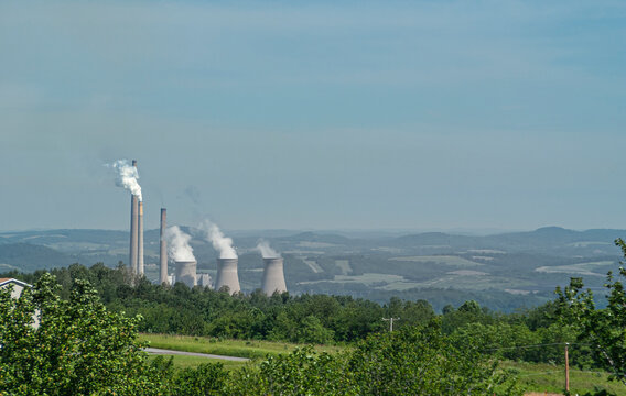 Johnstown, PA, USA - June 12, 2008: Mixed coal and nuclear power plant in Appalachian wilderness under light blue sky. Chimneys and cool towers spew white vapor clouds. Green foliage up front.