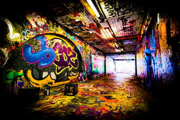 An underground pedestrian tunnel that has lots of graffiti on the walls, as well as a few cans of spray paint lying around.