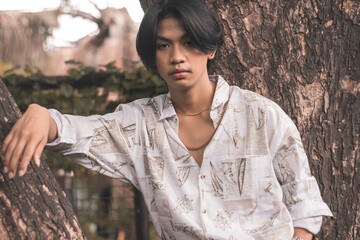 A young Filipino guy in a loose polo shirt and middle part straight hairstyle posing by a tree at a...