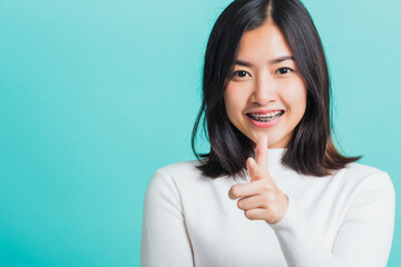 Young beautiful Asian woman smiling point finger at you with a confident expression, Portrait female pointing finger gesture towards you, studio shot isolated on a blue background