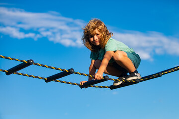 Cute boy climbs up the ladder on the playground. Child climbs up the ladder against the blue sky. Beautiful smiling cute boy on a playground. Kids activities.