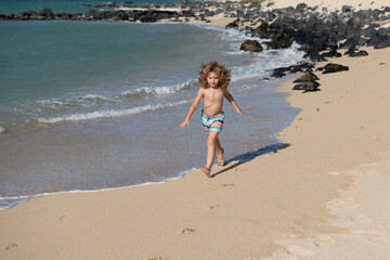Happy kid boy have fun on tropical sea beach. Funny child run with splashes by water pool along surf edge. Kids activity on summer holiday.