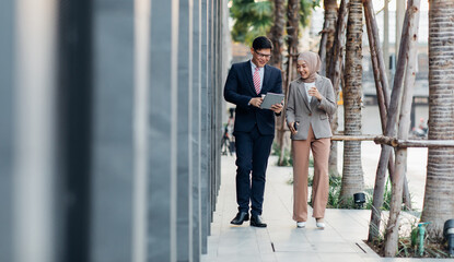 Young business muslim woman and business man colleague walking and discussing outside the office. Woman is holding a coffee and businessman is holding a digital tablet outdoor. - obrazy, fototapety, plakaty