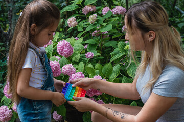 A girl and a mother click on a colored rainbow silicone anti-stress toy Push pop bubble. A child and his mother playing with a pop-fidget on a green background in a summer garden.