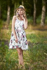 A young slender woman walks in the park on a summer day in a light summer sundress with a floral print in a hat . Long legs with heels.