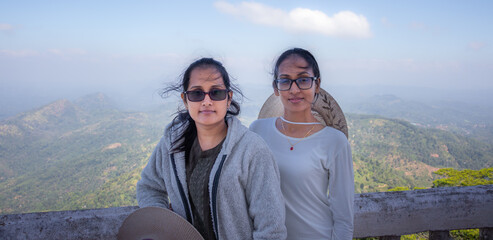 Two friends facing the camera in front of the breathtaking mountain range background of the distance. Spectacular viewpoint of Ambuluwawa tower.