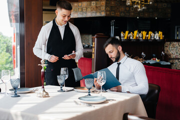 A young businessman in a fine restaurant examines the menu and makes an order to a young waiter in a stylish apron. Customer service.