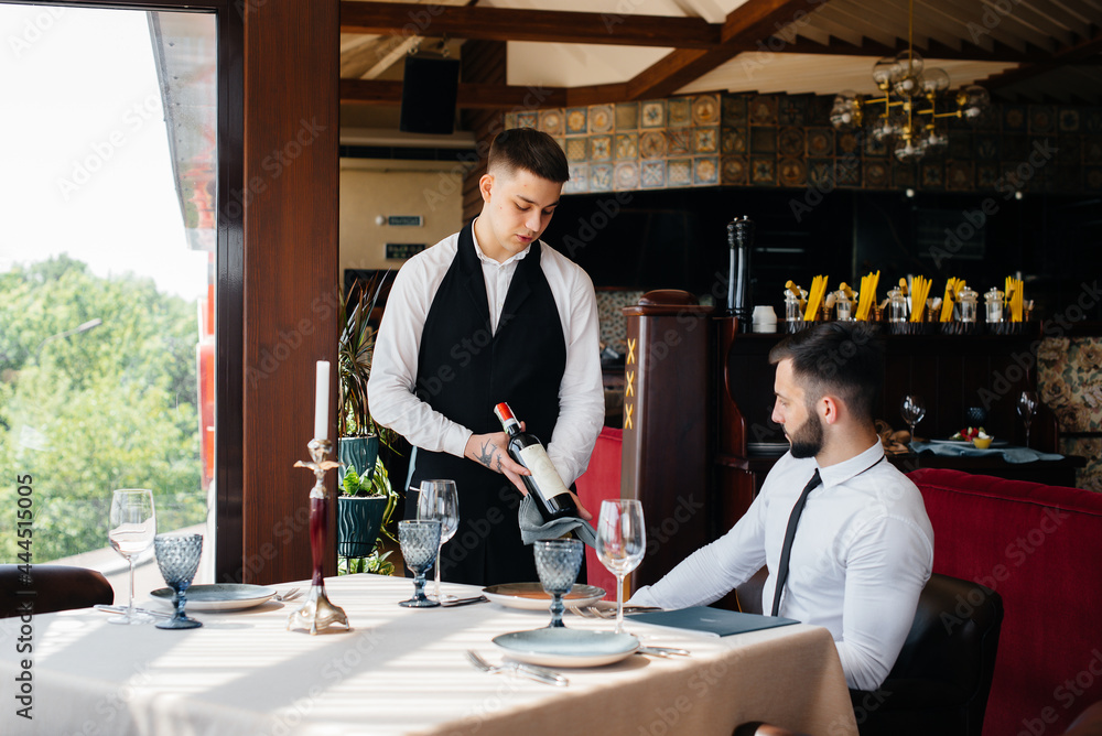 Wall mural A young waiter in a stylish apron demonstrates and offers a fine wine to a client in a restaurant. Customer service.