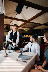 A young waiter in a stylish apron demonstrates and offers a fine wine to a beautiful couple in a restaurant. Customer service.