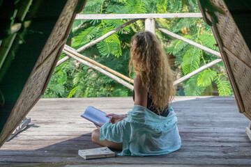 oung woman with curly long blonde hair reading book on balcony of tropical bungalow