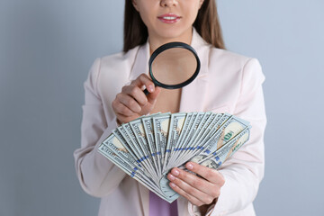 Expert authenticating 100 dollar banknotes with magnifying glass on light grey background, closeup....
