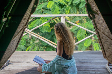 Young woman with curly long blonde hair reading book on balcony of tropical bungalow