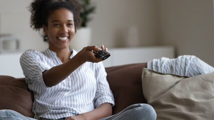 Close up smiling African American woman holding tv controller, switching channels, relaxing on cozy...