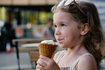 Toddler child eating cone ice cream outside near cafe. Cute blonde Caucasian girl licking frozen summer food on the street. Dirty mouth, happy kid.