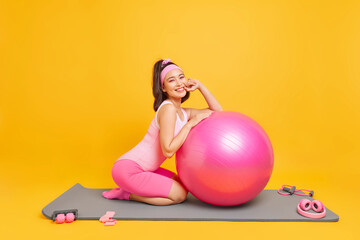 Fototapeta na wymiar Indoor shot of happy Asian sportswoman does exercising with fitness ball dressed in activewear leads healthy active lifestyle uses sport equipment isolated over yellow background. Athletic female