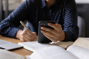 Female student using online learning app on smartphone, consulting online article, watching webinar and writing notes in notebook, doing study research. Hand of woman holding mobile phone, close up