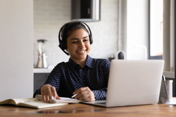 Happy Indian student in headphones with microphone attending virtual training class, watching webinar, taking notes and smiling. Employee working from home, talking to client via video call on laptop
