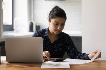 Focused millennial Indian woman counting budget, checking financial documents with receipts, using...