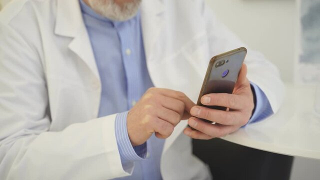 Cropped male Doctors in casual wear sit using smartphone at home, thinking, indoors. Close-up photo of hands, mobile phone.