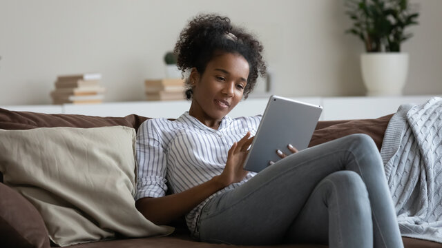 Smiling African American woman using tablet, relaxing on cozy couch at home, positive attractive young female looking at gadget screen, typing writing message in social network, chatting online